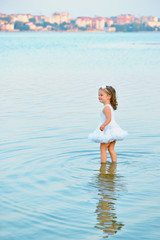 Adorable little toddler girl wearing beautiful white dress by a lake or sea on the beach in warm and sunny summer day