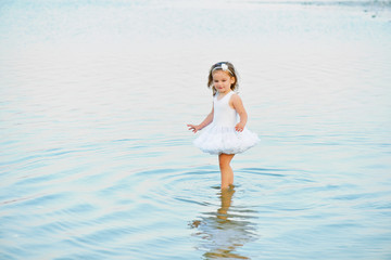 Fototapeta na wymiar Adorable little toddler girl wearing beautiful white dress by a lake or sea on the beach in warm and sunny summer day