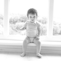 Cute baby at home in white room is sitting near window. The beautiful baby could be a boy or girl and is wearing body suit. Baby with curly hear is looking at camera. Black and white square photo.