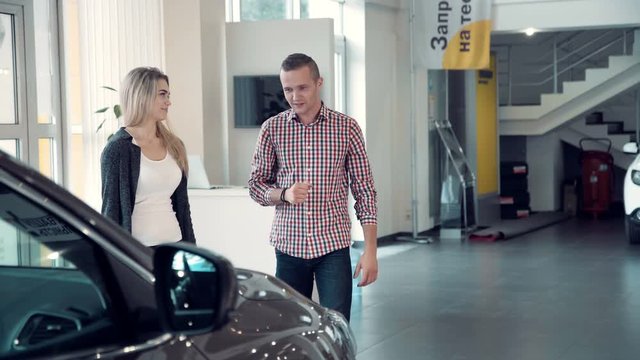 Young Salesman Delivering Sales Pitch to Couple Buying New Vehicle inside Car Dealership While Standing in front of Shiny New Model of car or electrocar