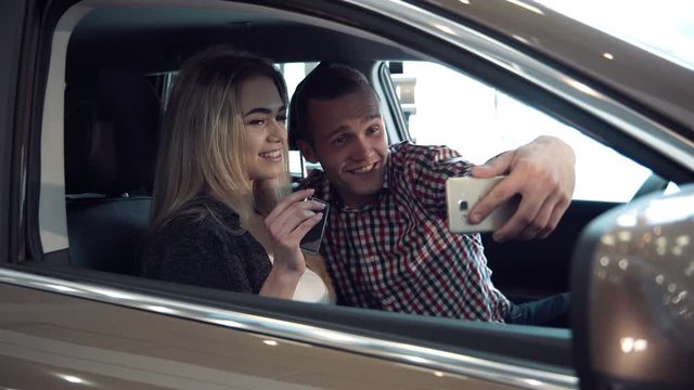 Excited young couple making a decision to purchase a new car sitting inside in a motor showroom checking it out for ownership and make selfie picture using smartphone.