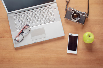  Overhead of modern comfort work place. Different objects on wooden background. Items include camera, glasses, laptop, smartphone and apple