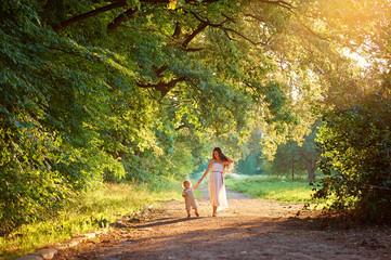Young mother and her little cute son walking in the park at sunset holding hands.