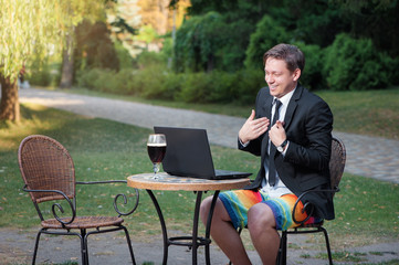 Work and relax. Online conference. Businessman dressed in suit and shorts working with laptop, talking by skype at the park cafe outdoors - obrazy, fototapety, plakaty