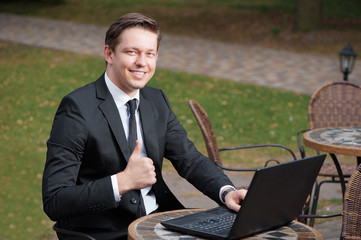 Good job! Happy young businessman in suit and tie showing his thumb up and smiling while working on laptop at cafe outdoors