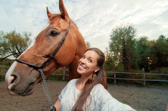 Funny selfie with my friend! Attractive smiling young woman holding camera and making selfie with her horse outdoors