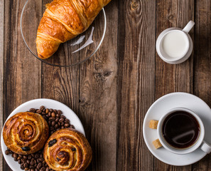 Coffee cup with milk jug, croissant and cinnabons on wooden background.