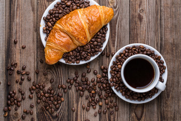 Breakfast coffee - cup and croissant with coffee beans on wooden background.