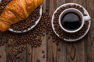 Breakfast coffee - cup and croissant with coffee beans on wooden background.
