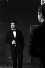 Young handsome stylish man in white jacket and black formal suit with bow-tie standing in interior and posing against mirror
