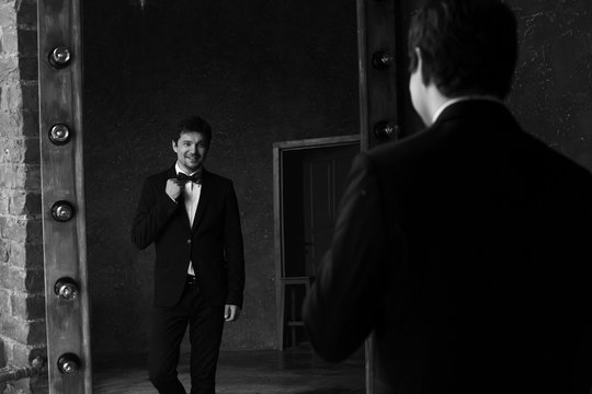 Young Handsome Stylish Man In White Jacket And Black Formal Suit With Bow-tie Standing In Interior And Posing Against Mirror