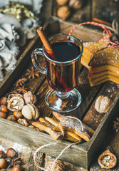 Glass of mulled wine in wooden tray with Christmas decoration toys, gingerbread cookies, nuts, cinnamon, anise and pine cones over rustic wooden background, selective focus, vertical composition