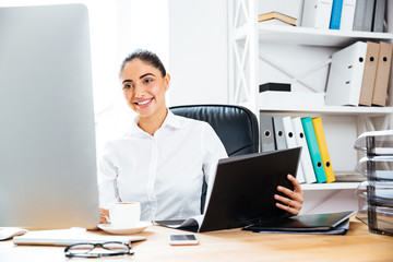 Fototapeta premium Smiling charming businesswoman holding documents and looking at computer screen