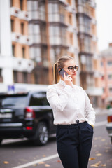 Business stylish girl with a mobile phone and glasses in the street in the city center on the background of cars and houses