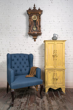 Still life of vintage blue armchair, yellow cupboard, pendulum clock and orange ornate scarf in studio