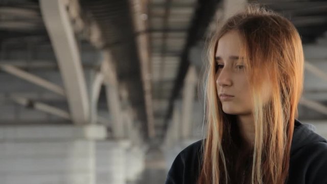 A teenage girl in a hoody with the loose multicolored hair spreading in the wind under the unfocused bridge pillars. The perspective receding into the distance between the bridge pillars.