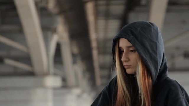 Skater style teenage girl in a hoody with the hood on and loose multicolored hair against the unfocused bridge pillar perspective receding into the distance.