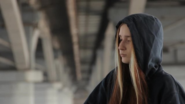 A teenage girl in a hoody with the hood on and loose multicolored hair against the unfocused bridge pillar perspective receding into the distance. Thinking deeply, taking the hood off.