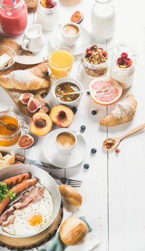 Fried egg with sausages and bacon, bread, croissants with jam and butter, fruits, smoothie, orange juice, yogurt, granola with milk and coffee on white wooden background. Selective focus, copy space