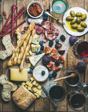 Wine and snack set with various wines in glasses, meat variety, bread, green olives, sun-dried tomatoes, figs, nuts and berries on wax paper over rustic wooden background, top view