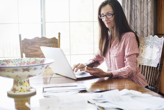 Senior woman with long brown hair sitting at a table, using a laptop computer, wearing reading glasses.
