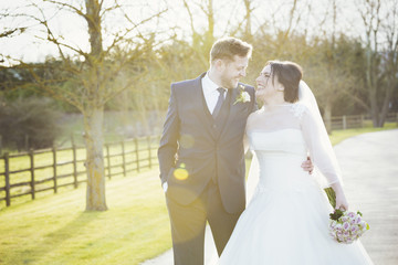 A bride and groom on their wedding day walking arm in arm down a path in the sunshine laughing. 
