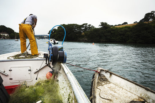 A fisherman in waders on a boat, hauling up the anchor.