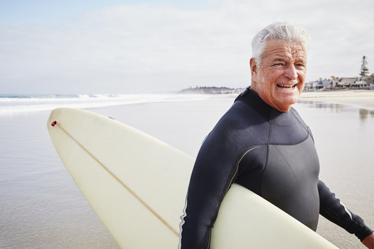 Smiling senior man standing on a beach, wearing a wetsuit and carrying a surfboard.