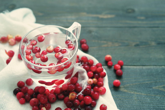 Fresh berries of a forest red cranberry in transparent glass with water among the scattered berries on a white linen cloth on a wooden surface of a table