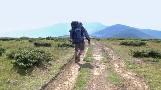 Man tourist with a big backpack goes on the grass against mountains