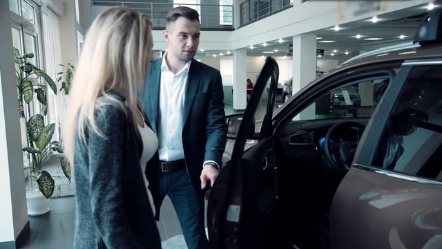 Salesman presenting a new electric or petrolium car to a female customer in a motor showroom. He tells about all privileges of owners of electric cars or just clients of their automobile center 4K