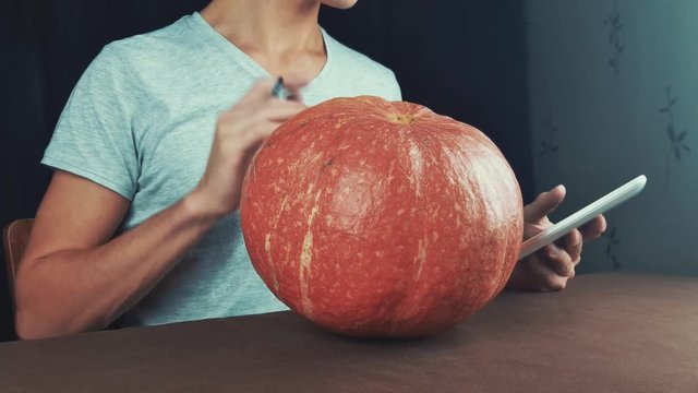 Guy in white shirt use picture on tablet computer as reference and draw on pumpkin jack o lantern 4k