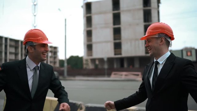 4k. UltraHD. Two businessmen architect in suits funny high five both arms than hit his helmets. They looks very happy of finishing project. Frends in business concept.