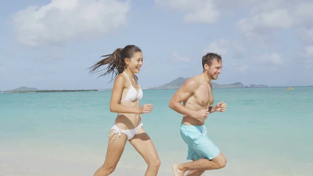 Happy couple having fun on beach vacation during summer holiday. Multiracial fit couple running together laughing in the sun. Young adults in shape carefree feeling good in their body.