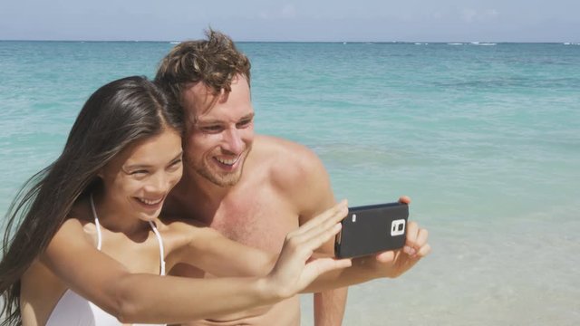Young multicultural happy couple taking selfie (self portrait) on beach using smartphone mobile cell phone camera. Young happy Asian woman and Caucasian man. RED EPIC SLOW MOTION 96 FPS.