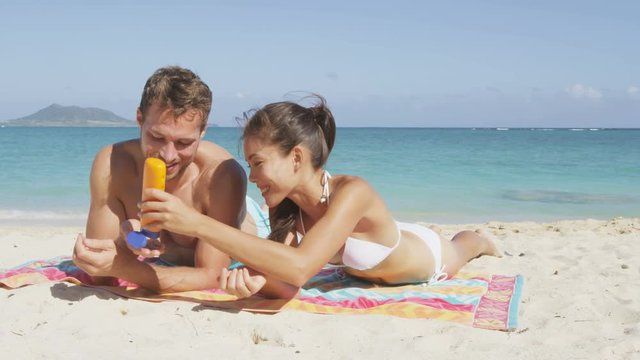 Woman helping man with sunscreen at beach. Happy couple sun tanning putting sunscreen suntan lotion having fun laughing and relaxing on holiday vacation travel. Multiracial Asian woman, Caucasian man.