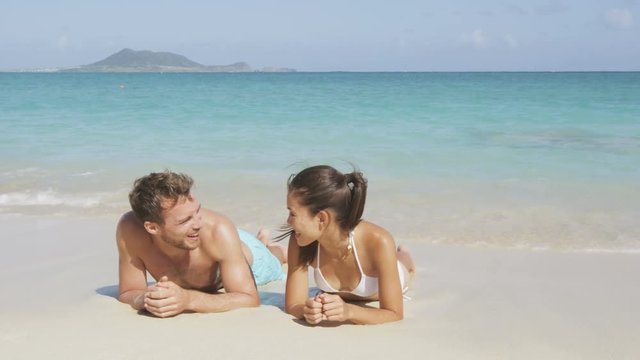 Couple in love relaxing on beach during vacation travel. Happy couple portrait talking together in a happy relationship sun tanning lying on the sand having fun in Hawaii, United States of America.