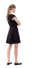 back view of standing young beautiful  woman.  girl  watching. Rear view people collection.  backside view of person.  Girl in black dress standing with his arms crossed.