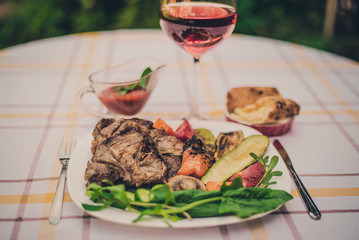Delicious juicy grilled steak, vegetables and mushrooms on the plate, surrounded by tomatoes, fresh herbs and red barbecue sauce. Glass of red wine and lavash.