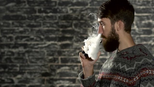 Young man with beard vaping an electronic cigarette. Vaper hipster smoke vaporizer in slow motion
