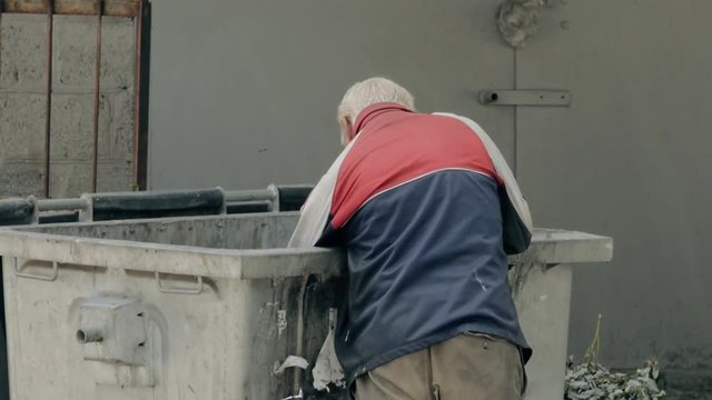 Homeless man rummaging through a dumpster. Hungry homeless man looking in garbage bins for food to eat on.