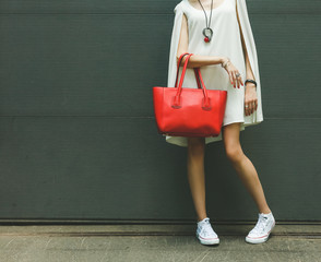 Fashionable beautiful big red handbag on the arm of the girl in a fashionable white dress, posing near the wall on a warm summer night. Warm color. - obrazy, fototapety, plakaty