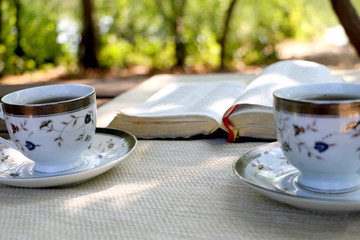  open book, two design cups of coffee on the wooden table in sunny day in the forest and mobile phone 
