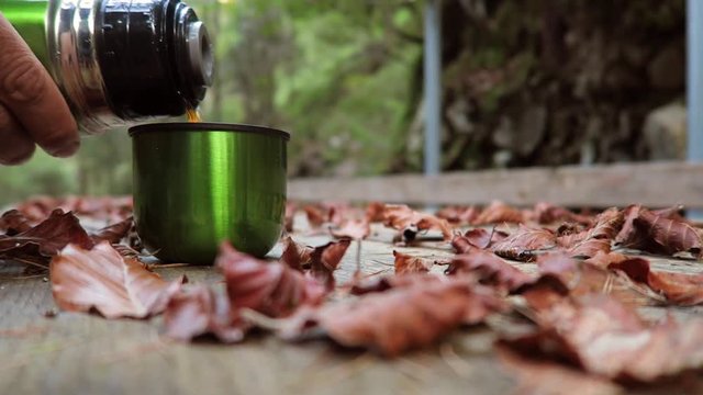 Man pours tea from a thermos into a mug on a wooden table among the autumn leaves. Wind blowing autumn leaves. Autumn mood background. Concept of the season, mood, hike, travel, time. 