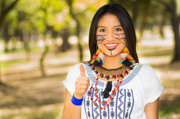 Beautiful Amazonian woman with indigenous facial paint and white traditional dress posing happily for camera in park environment, forest background