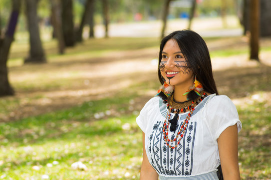 Beautiful Amazonian woman with indigenous facial paint and white traditional dress posing happily for camera in park environment, forest background