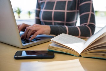 Woman using laptop with mobile phone and novel on table