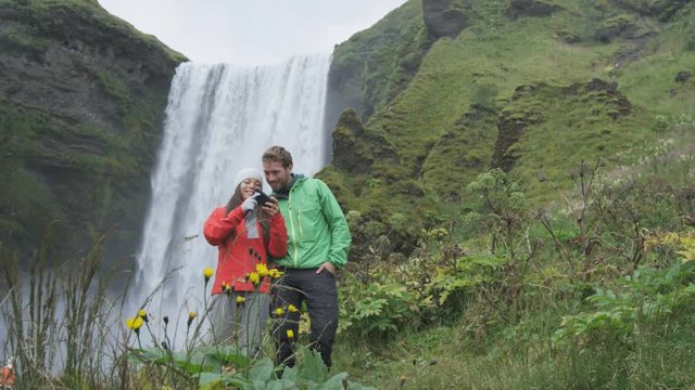 Selfie couple taking smartphone picture of waterfall outdoors in front of Skogafoss on Iceland. Couple visiting famous tourist attractions and landmarks in Icelandic nature landscape on ring road.
