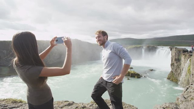 Couple taking photo with smartphone having fun by waterfall Godafoss on Iceland. Woman taking picture with smart phone camera on travel visiting tourist attractions and landmarks in Icelandic nature.