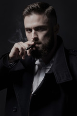 Attractive bearded man in a black coat smoking a cigar. He is stylish, strong and masculine, a cloud of cigar smoke rises above it. In his eyes shone a danger and sexuality.